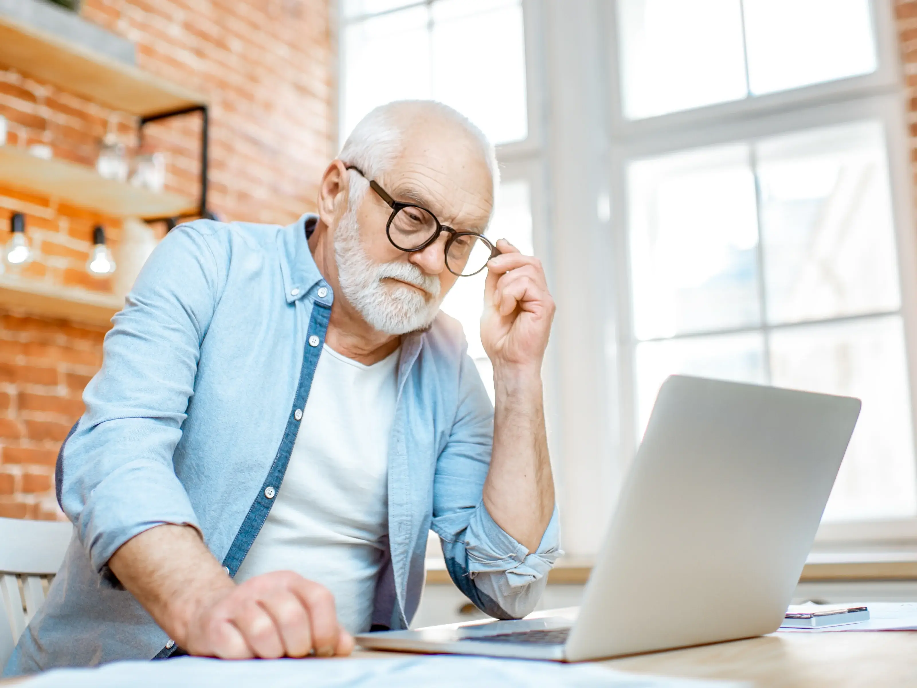 An elderly man with a beard and glasses sits at a desk with a laptop. Leaning slightly forward and adjusting his glasses, he focuses intently on the screen, as if analyzing a difficult problem or playing a challenging mahjong layout.