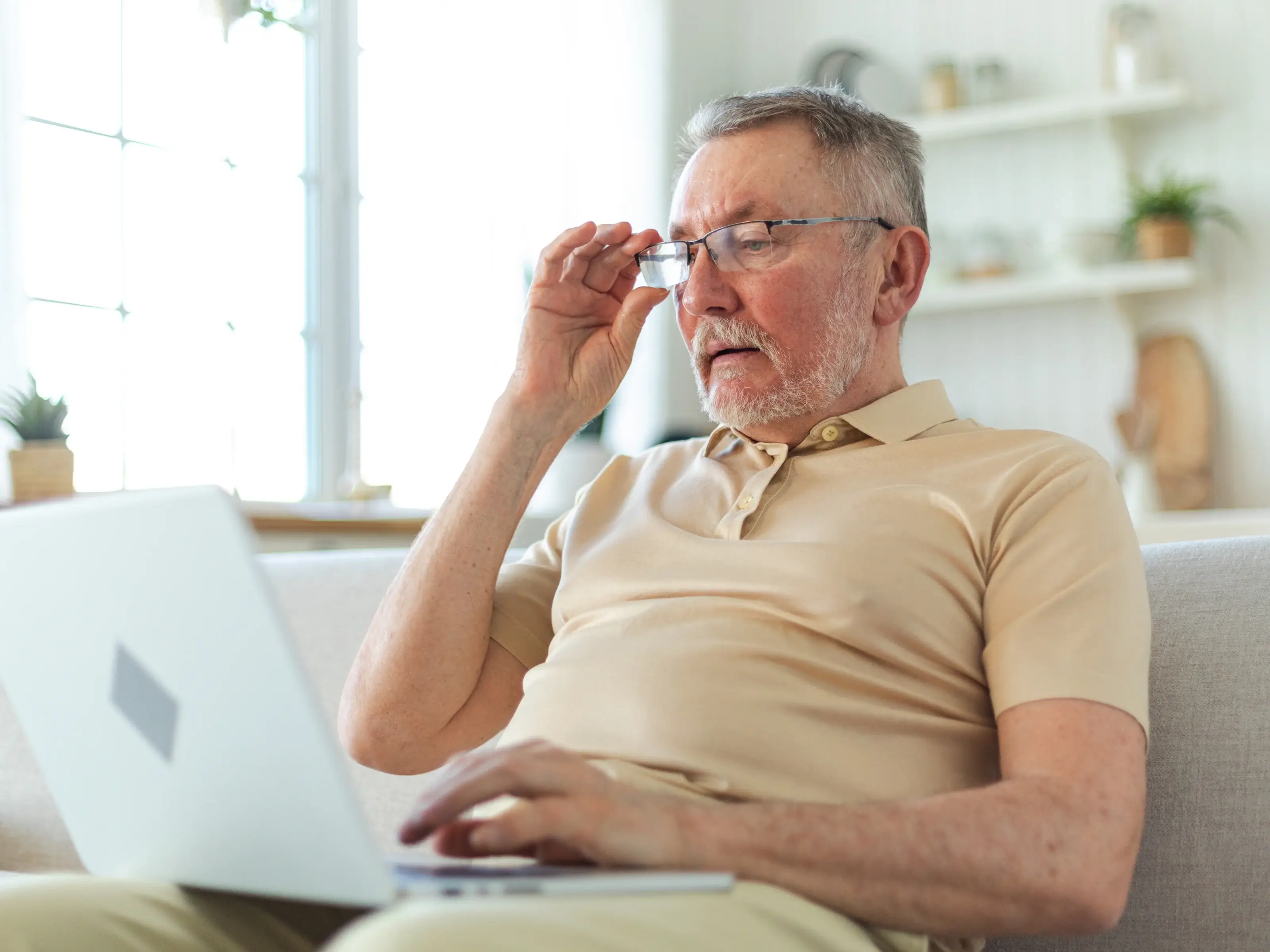 An elderly man wearing glasses and a light polo shirt sits on a couch with a laptop on his lap. He lifts his glasses and looks at the screen with a focused expression, as if solving a puzzle or playing a medium-difficulty mahjong game.