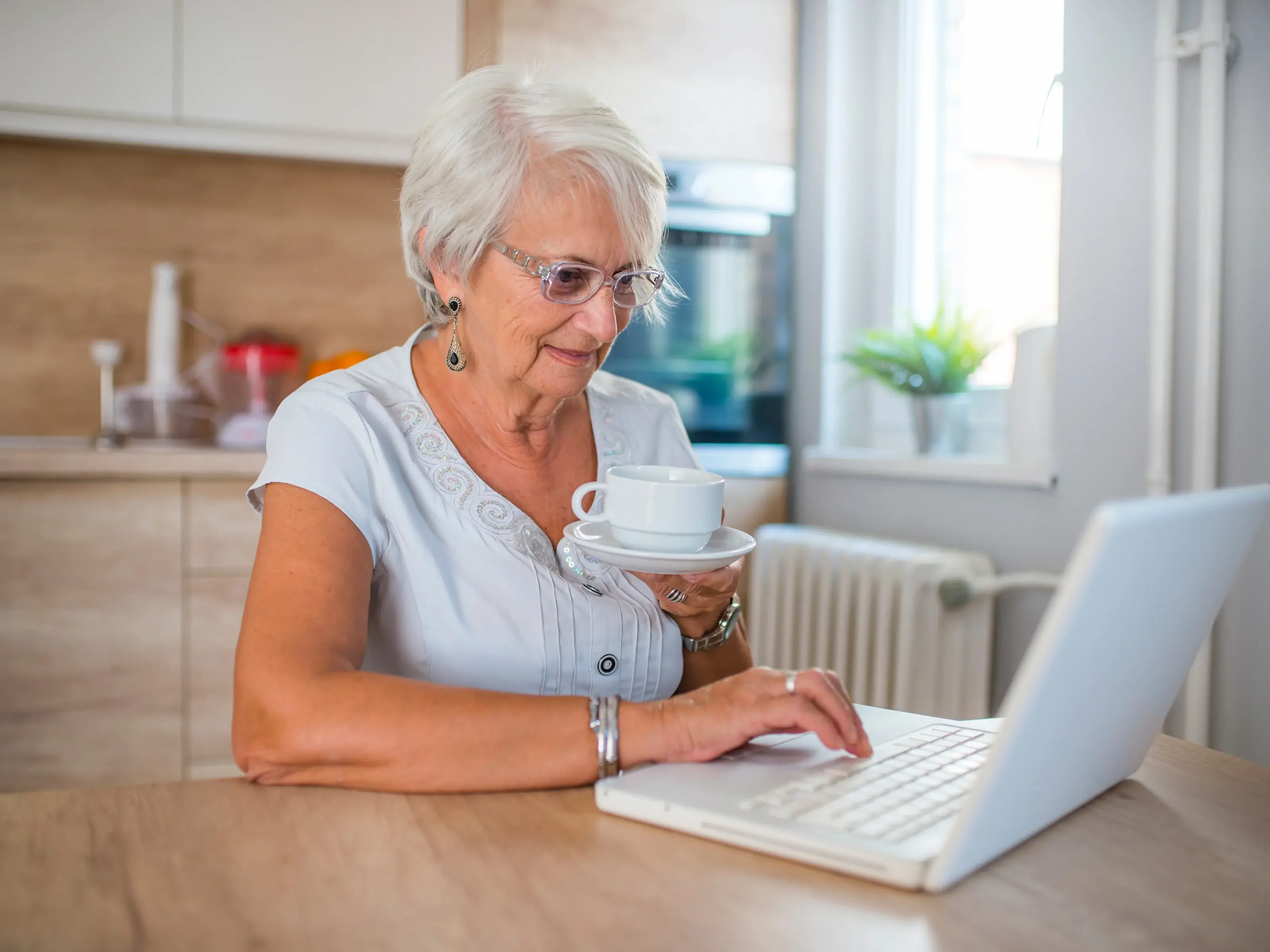 An elderly woman with gray hair and glasses sits at a kitchen table with a laptop. She holds a cup of tea or coffee and smiles while looking at the screen, creating a cozy and relaxing atmosphere.